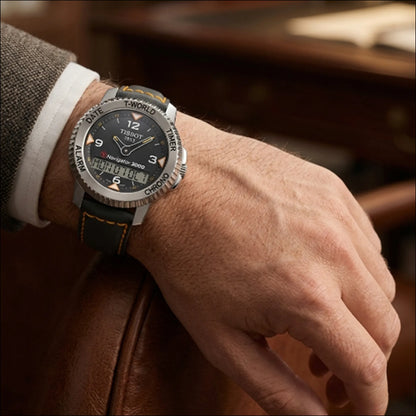 A close-up of a man's wrist wearing a Tissot T-Navigator watch with a grey dial, silver bezel, and black leather strap with orange & grey stitching, resting on a brown leather surface.